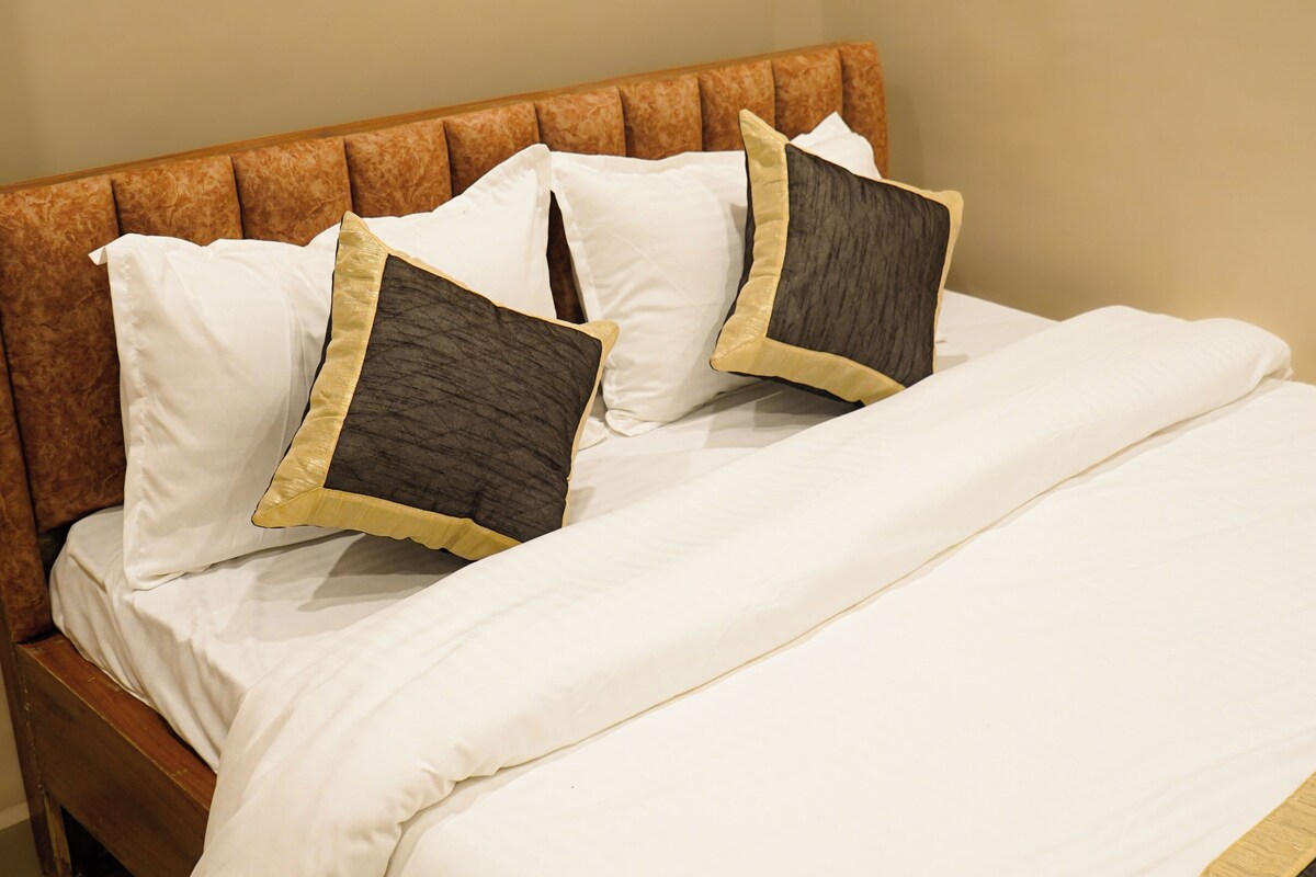 A bed with a wooden headboard, white bedding, and two pillows with a quilted pattern and a gold trim.