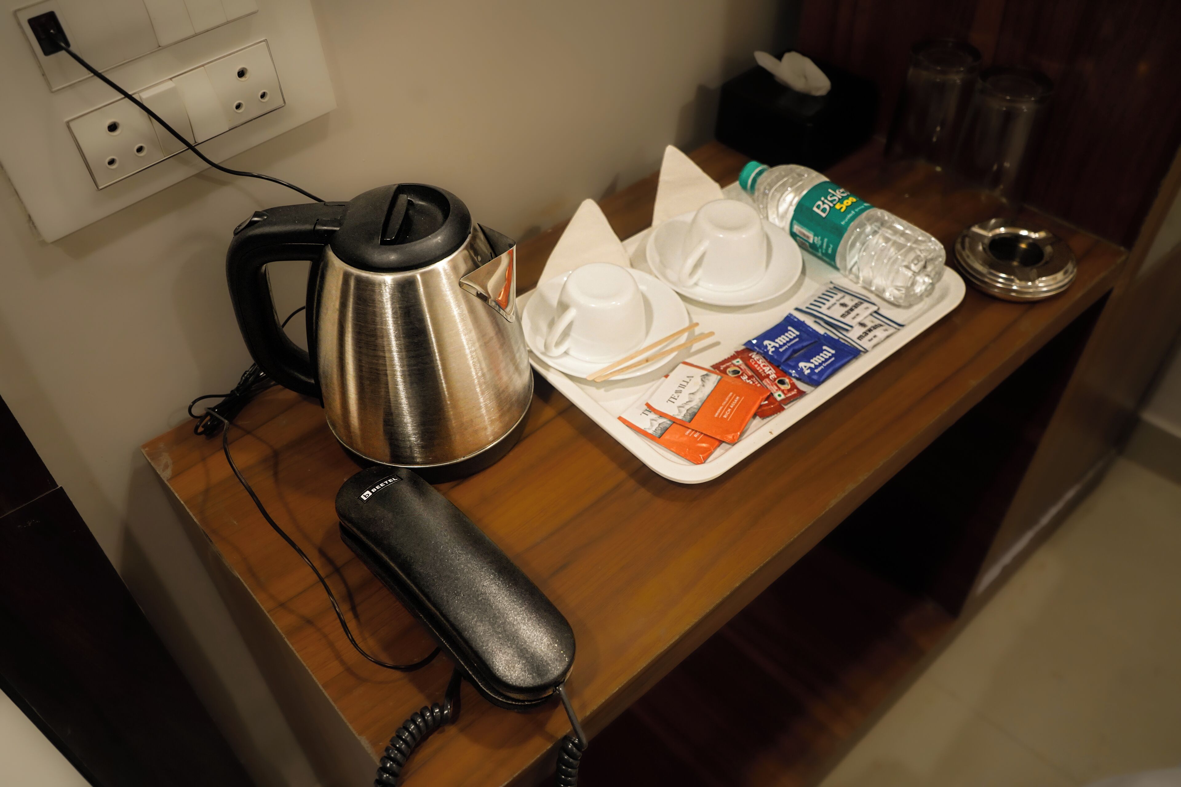 Ein Hotelzimmer mit einem Holztisch, einem Wasserkocher, einem Telefon und einer Ablage mit Tassen, Taschentüchern, einer Wasserflasche und einem Taschentuchpaket.