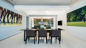 Un moderno comedor con una mesa y sillas de vidrio, una cocina al fondo y dos grandes cuadros colgados en la pared.