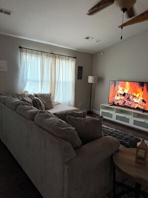 A living room with a sofa, a TV displaying a fire, and a ceiling fan. - Liberty INN- 4Beds- near Fort-Leonard-Wood (Waynesville)