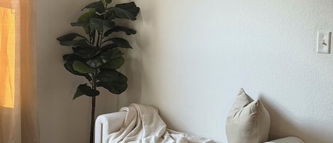 Una habitación con piso de madera oscura, un sofá blanco con una manta, una planta verde grande y una obra de arte enmarcada en la pared.