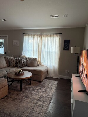 A living room with a sectional sofa, a round coffee table, a rug, a television, and a window with sheer curtains. - Liberty INN- 4Beds- near Fort-Leonard-Wood (Waynesville)