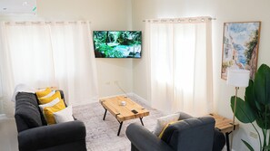 A living room with a TV mounted on the wall, a wooden coffee table, a sofa with cushions, and a painting on the wall. - Beautiful 3-bedroom house with AC in charming Ocho Rios (Ocho Rios)