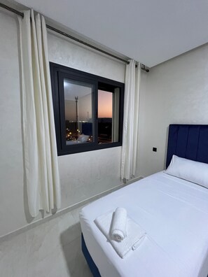 Habitación de hotel con cama, ventana con cortinas y vista a la ciudad al anochecer.