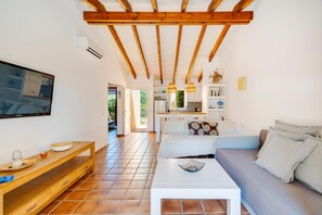 A modern living room with a wooden ceiling, a TV mounted on the wall, a sofa, a coffee table, and a kitchen visible through an open door. - Casa Massieu - Relax steps from the beach (Ciutadella de Menorca)
