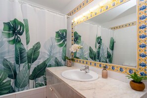A bathroom with a white sink, a mirror with a decorative frame, and a shower curtain with a tropical leaf pattern. - Casa Massieu - Relax steps from the beach (Ciutadella de Menorca)