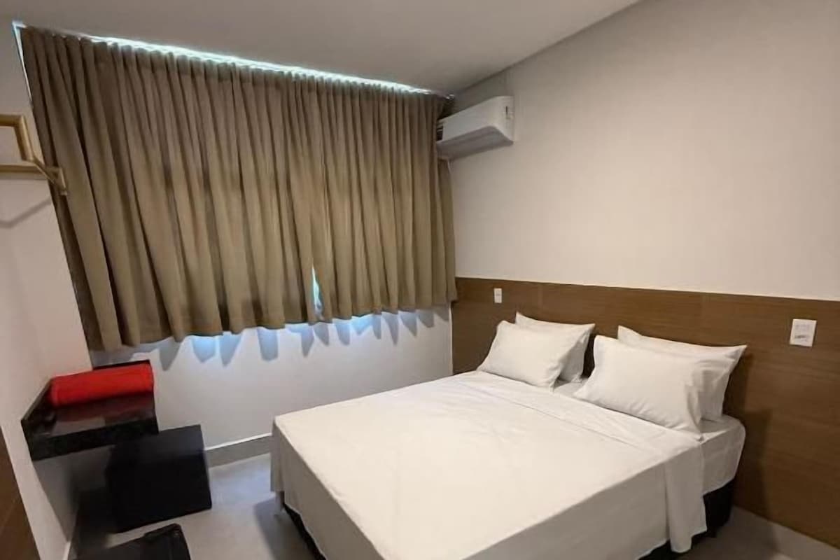 A hotel room with a bed, a bedside table, a chair, a red bench, and a window with curtains.