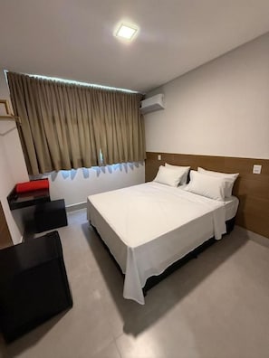 Une chambre d’hôtel avec un lit, une table de chevet, une chaise, un banc rouge et une fenêtre avec des rideaux.