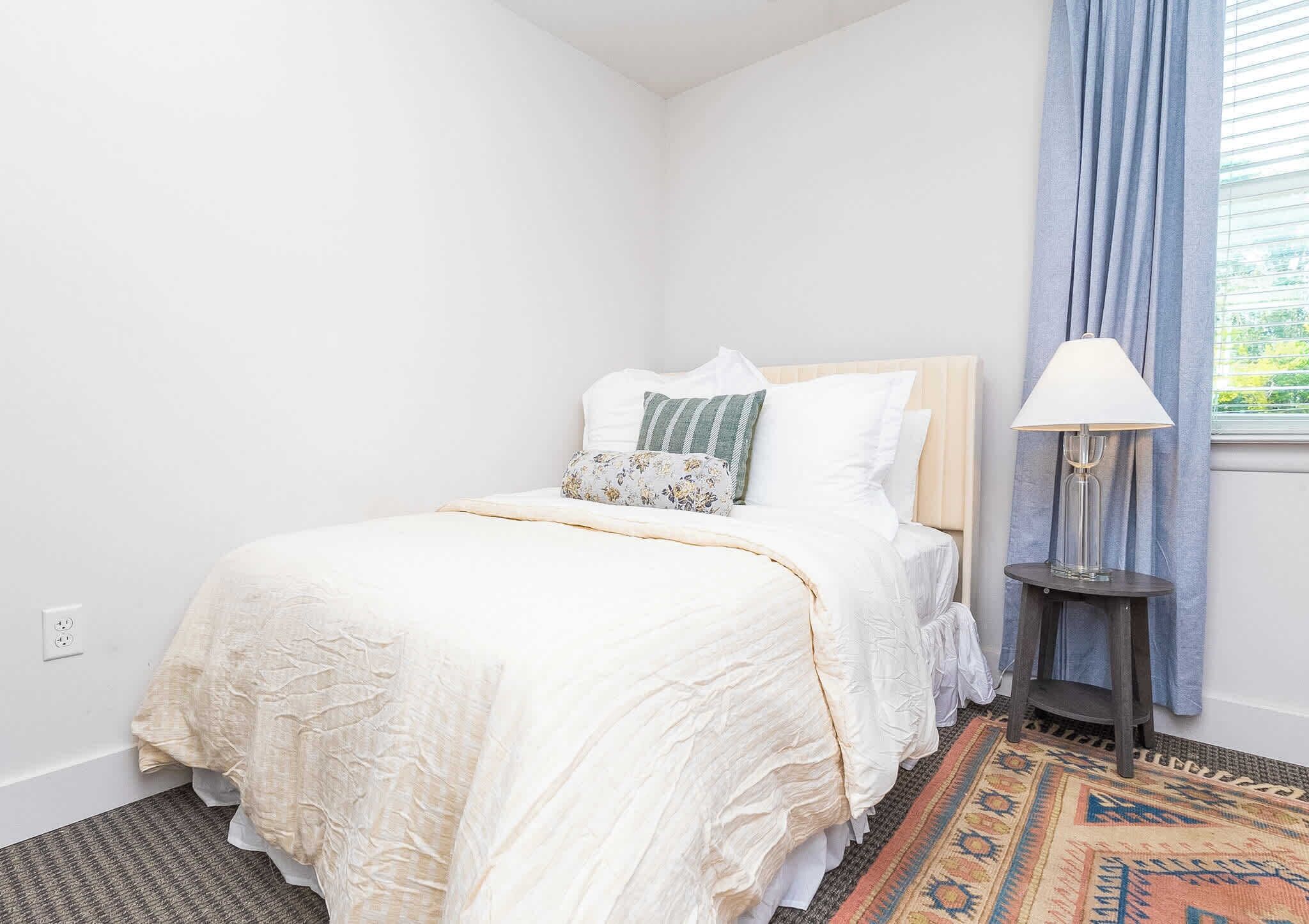 Un dormitorio con una cama, una mesita de noche, una lámpara y un ventanal con cortinas.