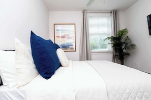 Ein Schlafzimmer mit einem Bett, Kissen, einem gerahmten Bild, einem Fenster mit Vorhängen und einer Topfpflanze.