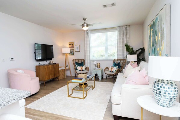 A modern living room with a flat-screen TV, a ceiling fan, and a large window with blinds.