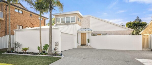 A modern two-story house with a garage, a covered driveway, and a landscaped front yard with palm trees and shrubs. - Casa En Norah | Coastal Escape (Norah Head)