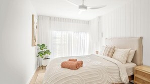 Een slaapkamer met een bed, een plafondventilator, een nachtkastje, een plant in pot en een raam met dunne gordijnen.