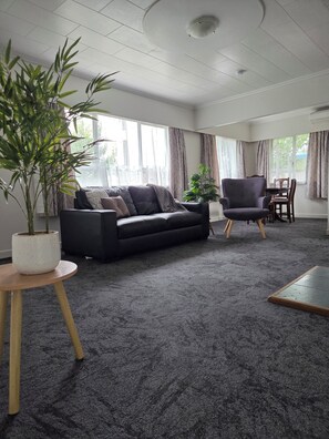 Una sala de estar con alfombra gris oscuro, un sofá negro, una mesa de centro de madera y dos plantas en macetas.