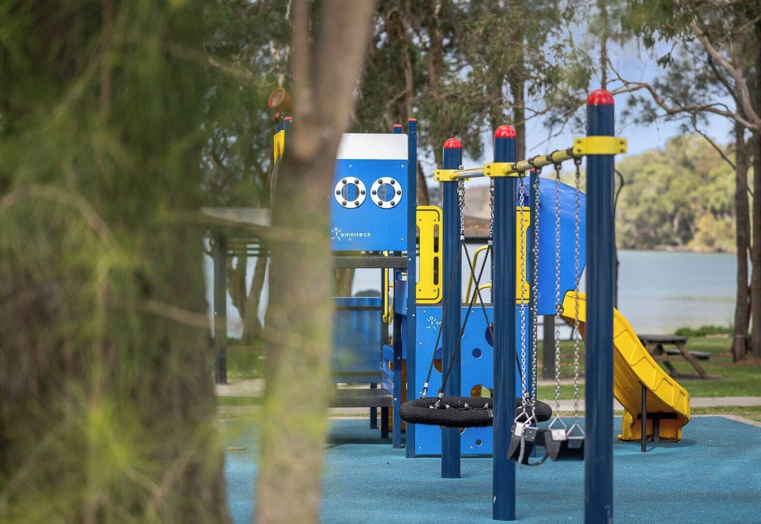 A playground with a blue and yellow play structure, a swing set, and a slide, surrounded by trees and a body of water.
