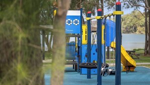 A playground with a blue and yellow play structure, a swing set, and a slide, surrounded by trees and a body of water.