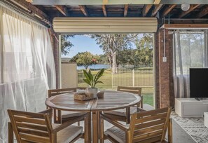 Un espacio exterior techado con comedor, mesa, sillas, televisor y vistas a un lago y árboles.