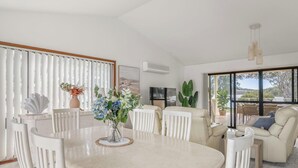 Ampla sala de estar com mesa de jantar, cadeiras brancas, sofá e uma grande janela com vista para o exterior.