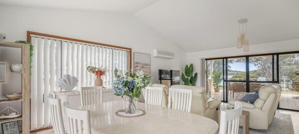 A spacious living area with a dining table, white chairs, a sofa, and a large window offering a view of the outdoors.