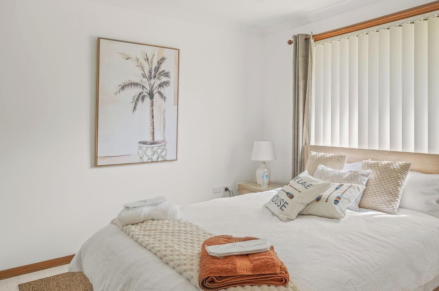 A hotel room with a bed, pillows, a nightstand, a lamp, a framed picture of a palm tree, and a window with curtains.