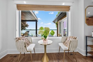 Una habitación con dos sillas frente a un ventanal que da a una masa de agua, una mesa redonda con un florero y un espejo en la pared.