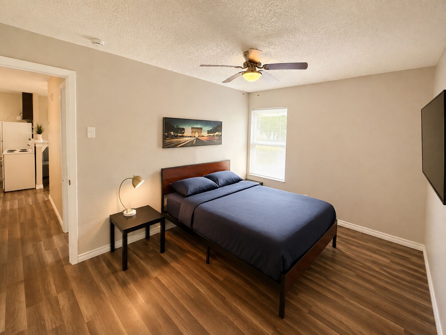 A bedroom with a bed, a nightstand, a ceiling fan, a window with blinds, and a door leading to a kitchen.
