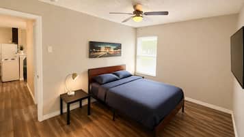 A bedroom with a bed, a nightstand, a ceiling fan, a window with blinds, and a door leading to a kitchen.