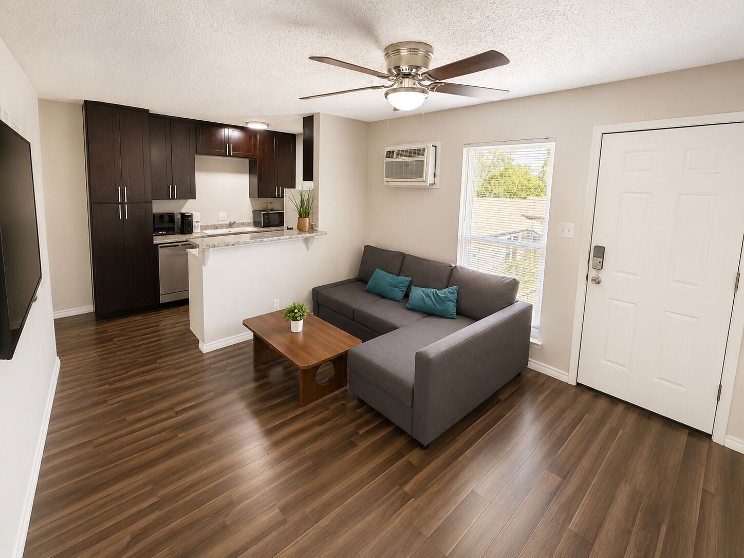 A modern apartment with a kitchenette, grey sofa, wooden flooring, and a ceiling fan.