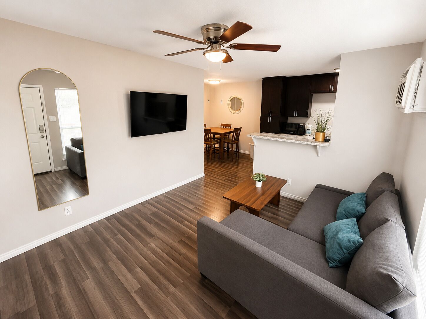 A modern living room with a flat-screen TV, a ceiling fan, a dining area with a table and chairs, and a kitchen with dark cabinets and a countertop.