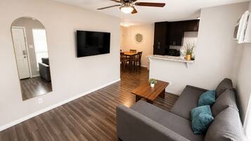A modern living room with a flat-screen TV, a ceiling fan, a dining area with a table and chairs, and a kitchen with dark cabinets and a countertop.