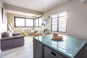 A modern living room with a glass coffee table, grey sofa, and a dining area with a glass table and chairs. - Arecibo 3BR | Beach Views | Jacuzzi (Arecibo)