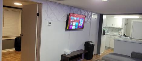 Ein modernes Wohnzimmer mit einem Wand-Fernseher, einem Holztisch, einem grauen Sofa und einem durchgehenden Durchgang zur Küche.