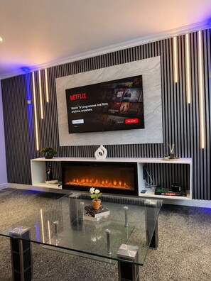 Ein modernes Wohnzimmer mit einem großen Flachbildfernseher an der Wand, einem Kamin darunter und einem Glastisch im Vordergrund.