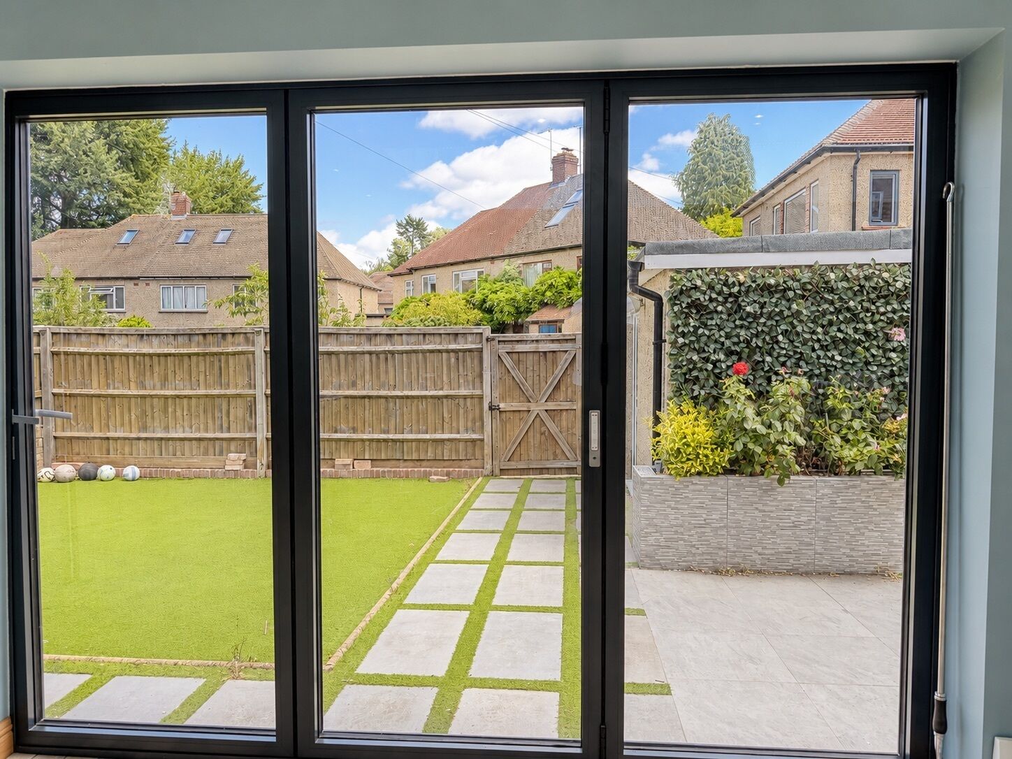 On aperçoit, à travers une porte-fenêtre coulissante, un jardin avec une clôture en bois, un chemin pavé et un mur végétal.