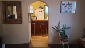 An interior hallway leading to a kitchen area with wooden stools, a potted plant, and a framed picture on the wall. - Charming Historic Las Cruces House & Farmers Market (Las Cruces)