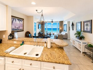 A kitchen with granite countertops, a white sink, and a dining area with a view of the ocean. - Portofino- Gulf Front with Beautiful Sunsets! (Escambia County)
