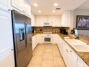 A modern kitchen with white cabinets, a stainless steel refrigerator, a microwave, an oven, and a granite countertop. - Portofino- Gulf Front with Beautiful Sunsets! (Escambia County)