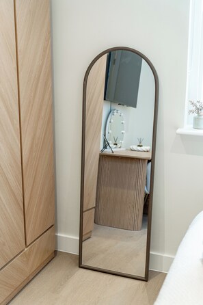 Ein Zimmer mit einem Holzschrank, einem Spiegel mit Holzrahmen, einem kleinen Schreibtisch ebenfalls mit Spiegel und einem Fenster mit Pflanze.