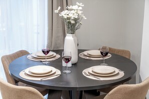 Una mesa de comedor redonda con platos blancos, cubiertos y dos copas de vino tinto. Un jarrón blanco con flores blancas está centrado en la mesa.