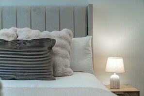 Ein ordentlich bezogenes Bett mit einem gestreiften Kissen und einem flauschigen Kissen, einer Nachttischlampe und einem Holz-Kopfschild.