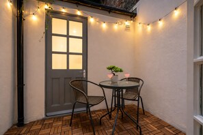 Un patio en esquina con una puerta oscura, dos sillas de mimbre, una mesa redonda y una pequeña planta en maceta.