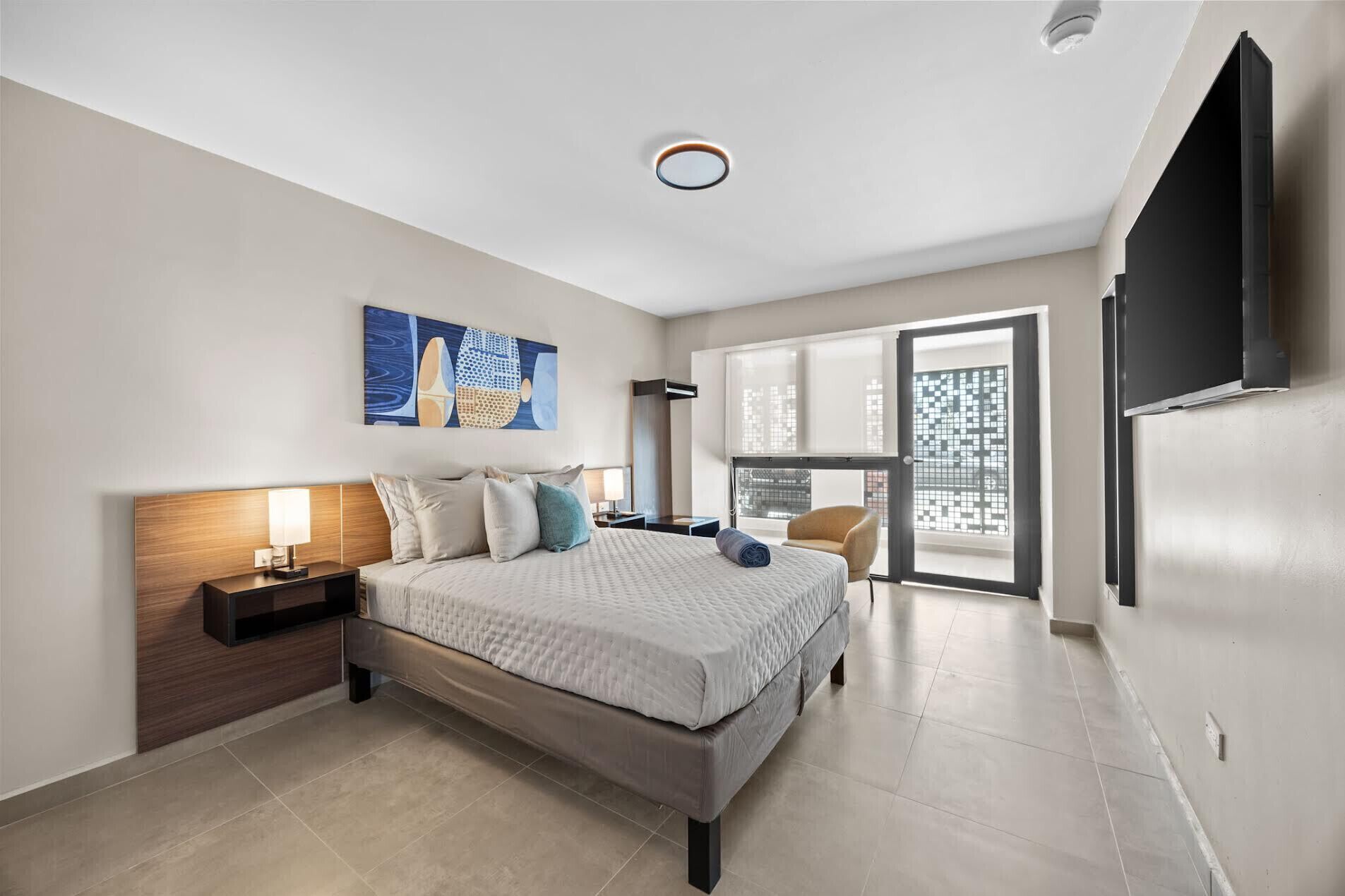 A modern hotel room with a large bed, a flat-screen TV, a painting, and a view of the city through the window.