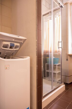 Un baño de hotel con cabina de ducha de vidrio, lavadora blanca y lavabo azul.