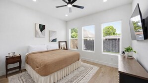 Quarto com cama, mesas de cabeceira, ventilador de teto, janelas e televisão.