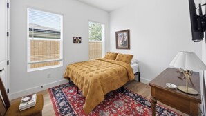 Quarto com cama, escrivaninha, cadeira, ventilador de teto e duas janelas com persianas.