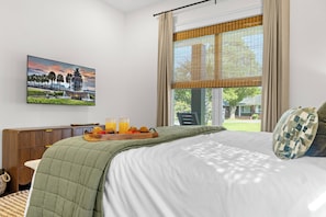 A hotel room with a bed, a wooden side table with a tray of fruit and juice, a painting on the wall, and a window with a view of the outdoors. - The Palmetto Haven House (North Charleston)