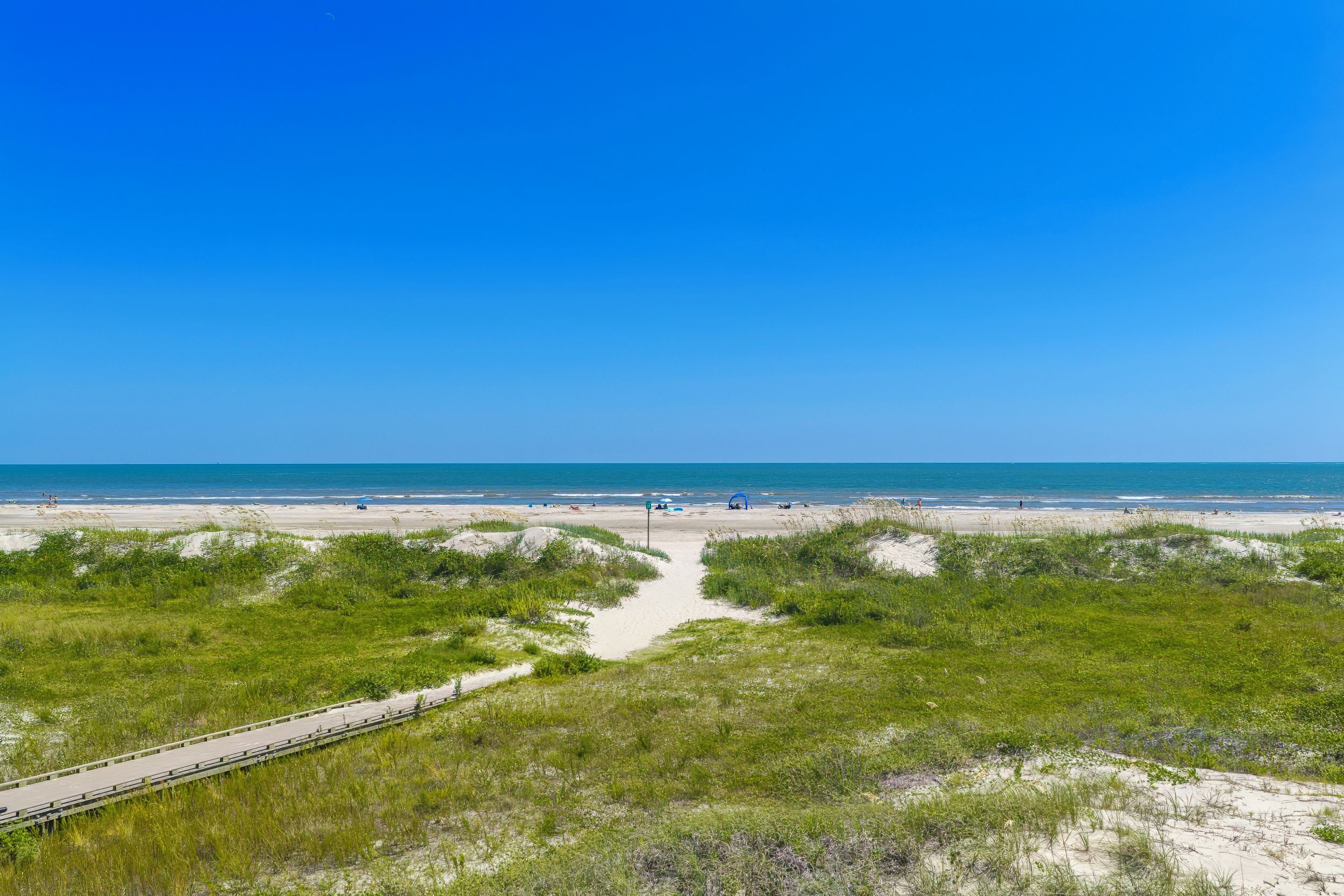 Una playa con un paseo marítimo de madera que conduce al agua, rodeada de dunas y vegetación.