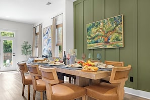 A dining area with a long table set for six, surrounded by chairs, with artwork on the walls and a view of the outdoors. - The Palmetto Haven House (North Charleston)