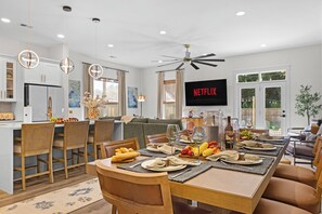 Una cocina moderna con armarios blancos, un comedor con mesa de madera dispuesta para una comida, y una sala de estar con sofá y televisor mostrando 'NETFLIX'.