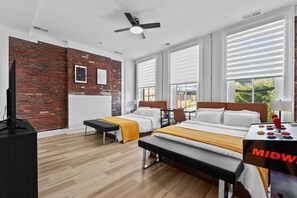 A hotel room with a brick wall, two beds, a desk, and a view of greenery. - Hot Tub | 6BR-4BA | Sleep 12+ | Game Rm | Fire Pit (Louisville)